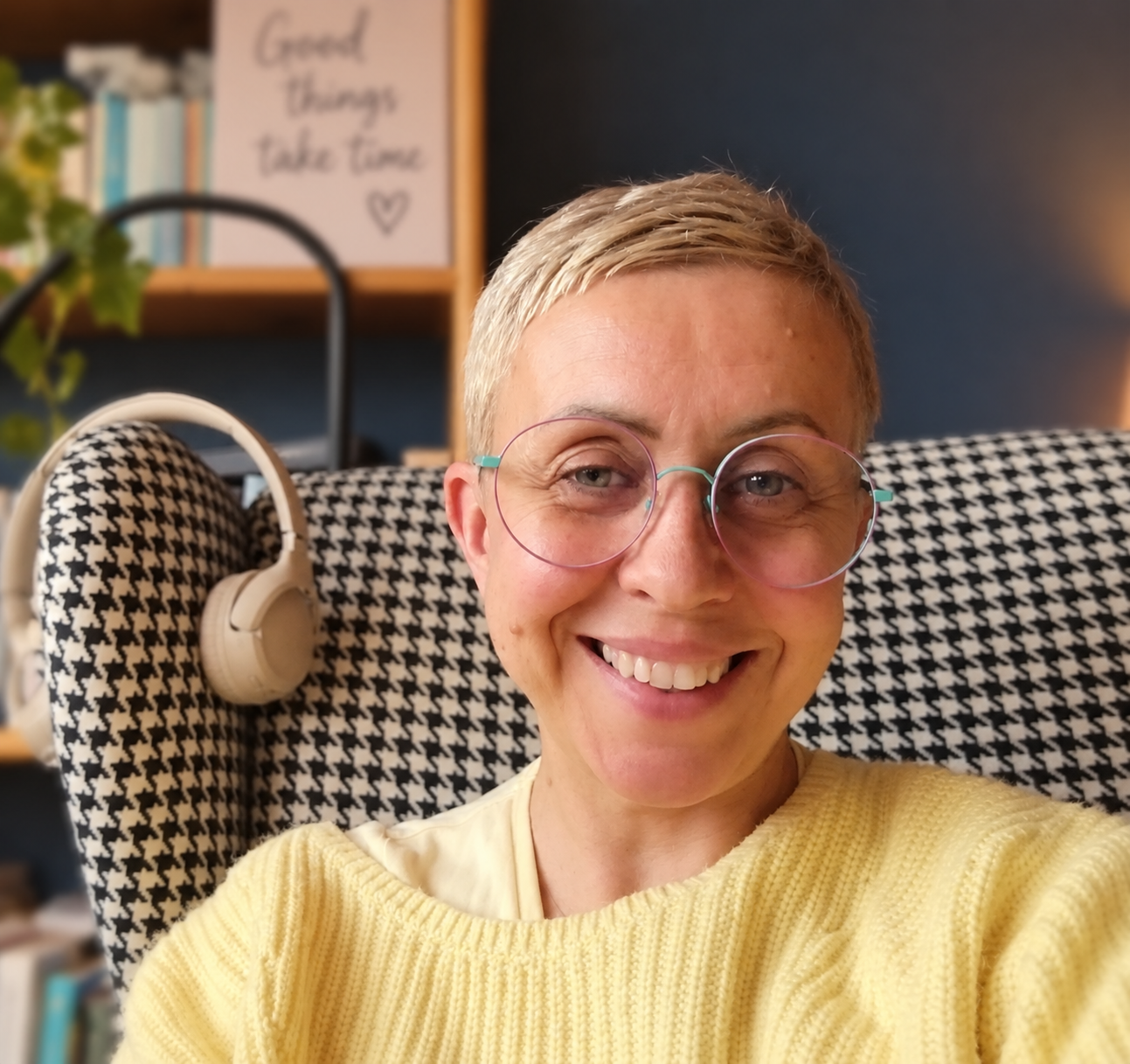 Karolina w żółtym swetrze w fotelu w pepitkę, obok biblioteczki i słuchawek